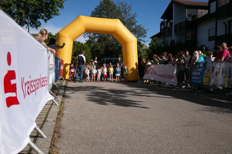 Blick entlang der Absperrung mit Sponsorenbannern zum Startbogen, unter dem sich Kinder aufstellen; Zuschauer säumen die Strecke.